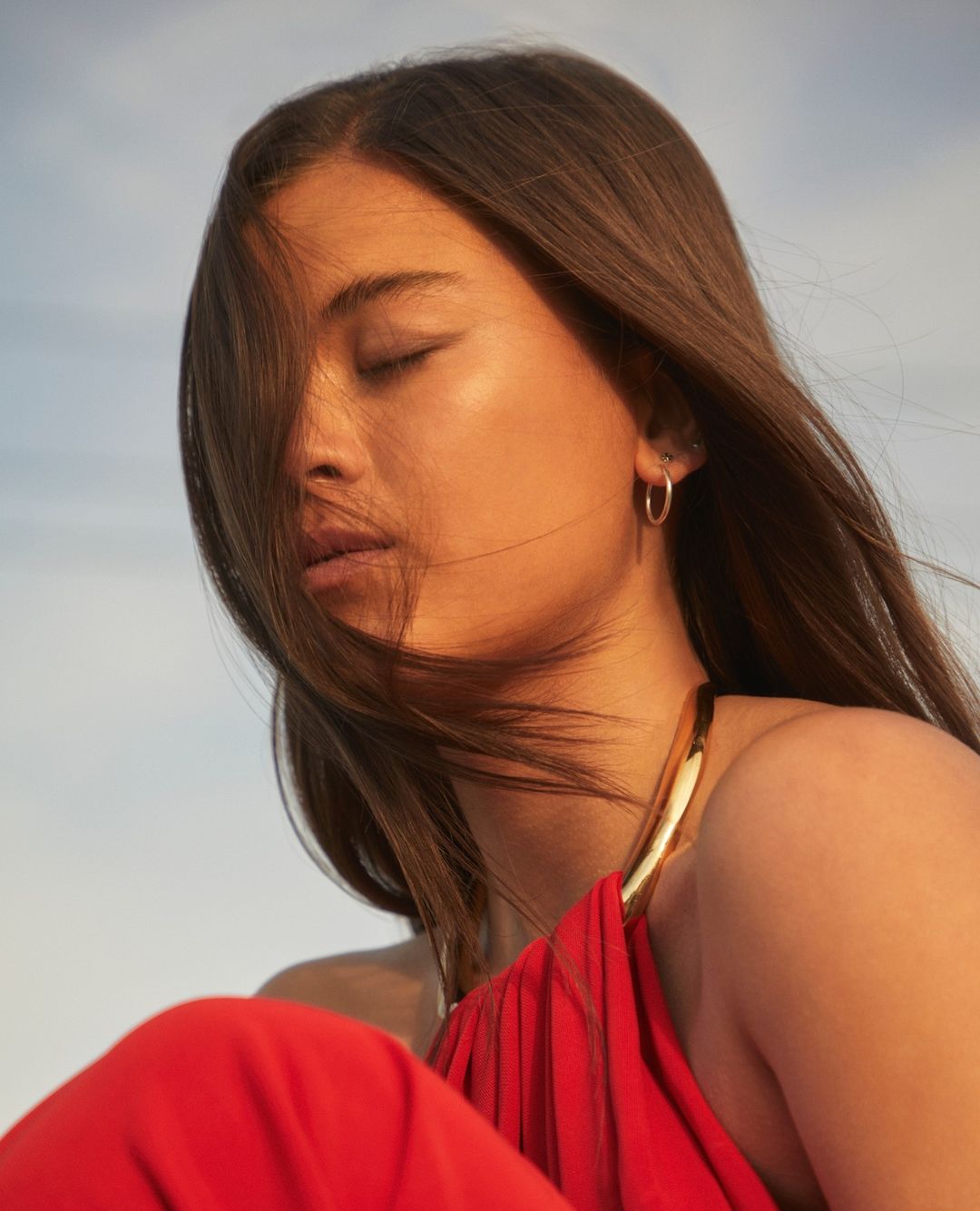 Close-up portrait of a woman with long brown hair wearing a red halter dress against a clear sky, photographed by Thomas Giddings.