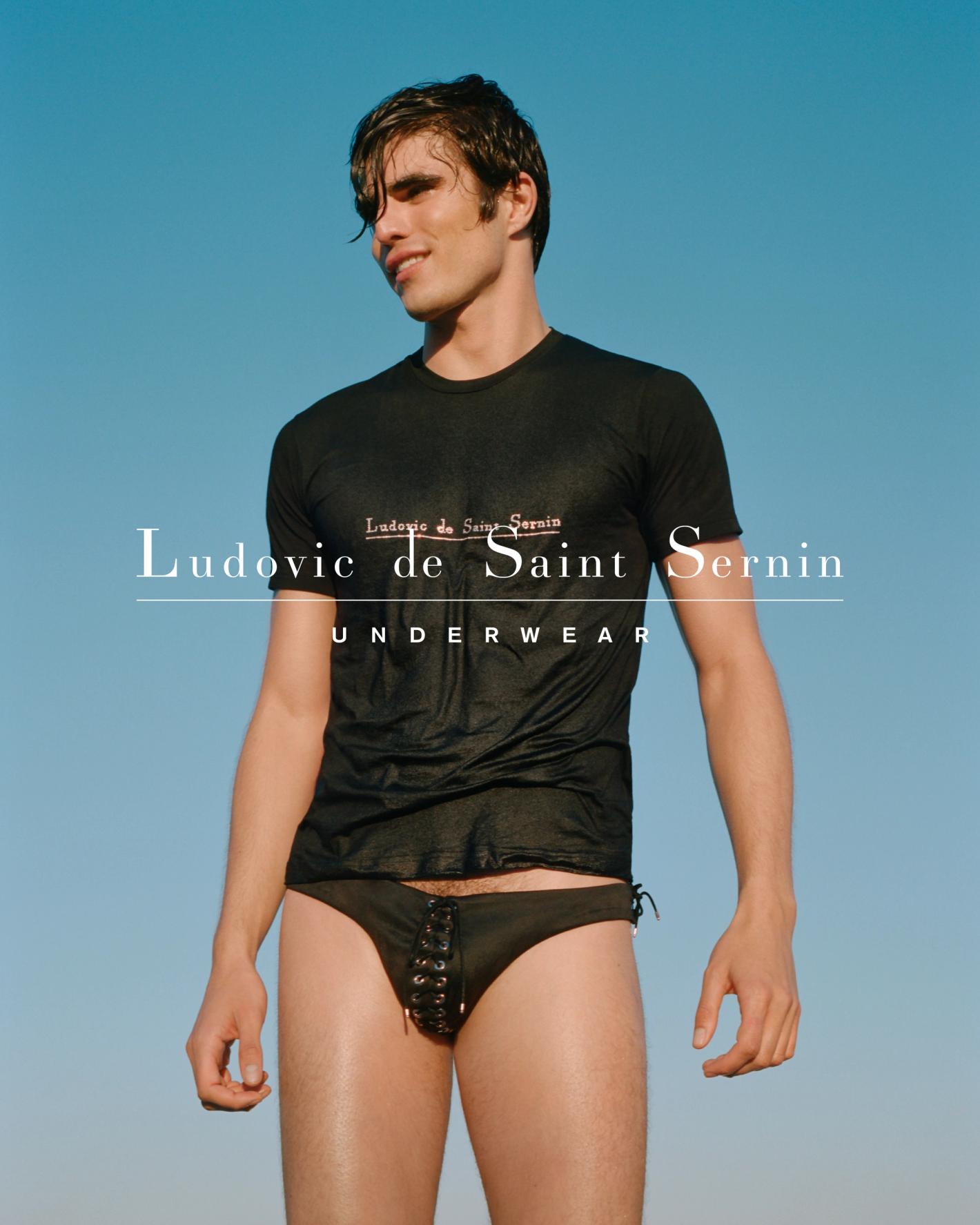 A male model poses against a blue sky, wearing a black t-shirt with logo and black laced swimwear bottoms, showcasing fashion and style.