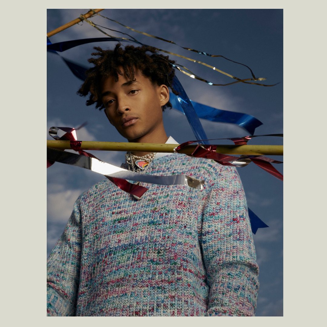 Young man with dreadlocks, wearing a multicolored sweater and necklace against a blue sky with metallic and fabric ribbons.