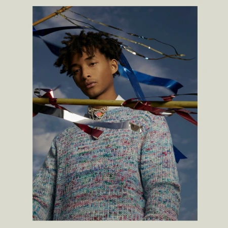 Young man with dreadlocks, wearing a multicolored sweater and necklace against a blue sky with metallic and fabric ribbons.