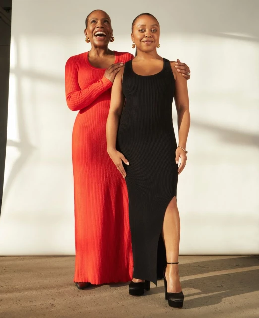 Two adult women in full-length ribbed dresses pose in a bright, indoor setting. The woman on the left wears an orange dress, the other a black dress.
