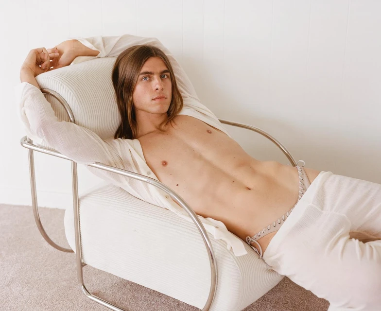 A light-skinned male model with long brown hair, lying partially undressed on a cream-colored chair in a photo studio setting.