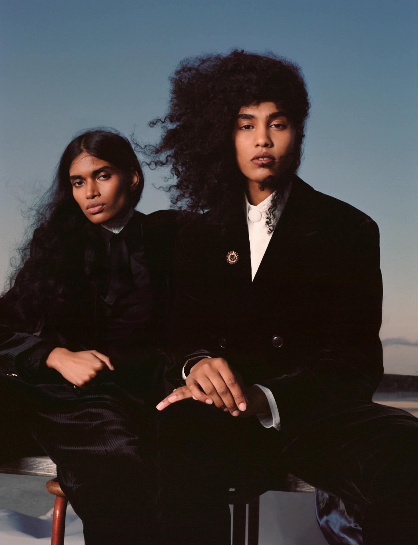 Two striking individuals with dark hair and dark jackets pose against a clear blue sky, creating a high-fashion, editorial vibe.
