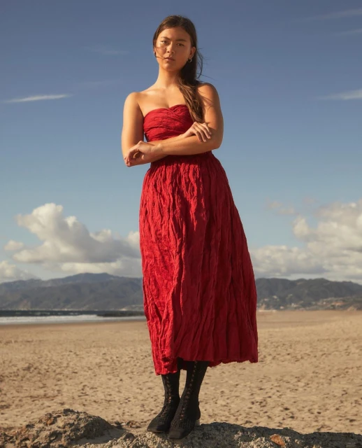A woman stands on a rocky beach in a red strapless dress and black lace-up boots, posing against a backdrop of mountains and a blue sky.