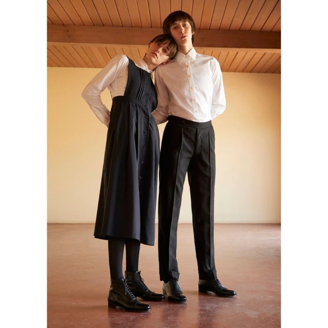 Two models, a female and a male, pose indoors wearing a white button-down shirts, black dress and trousers, paired with black boots.