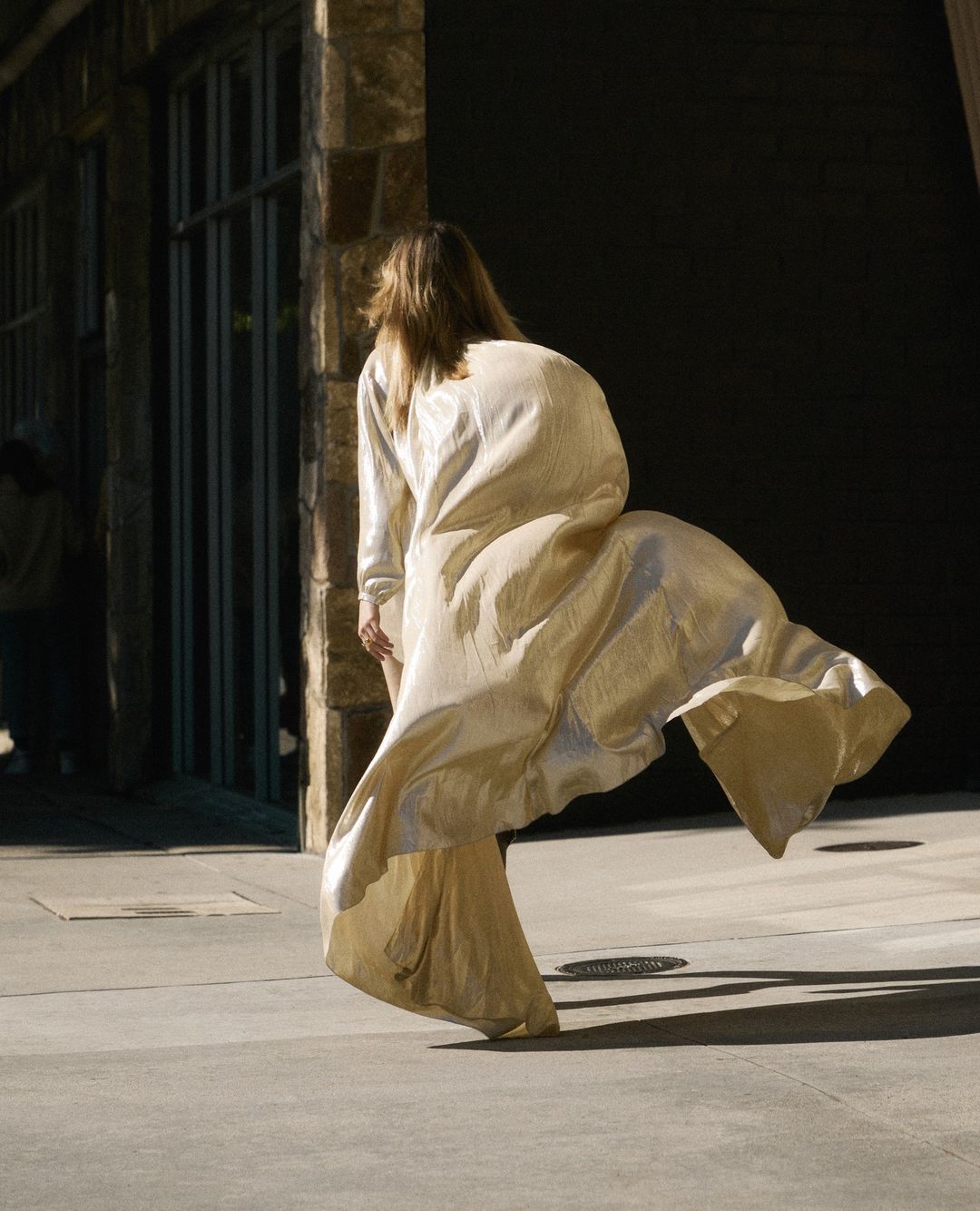 Adult female in flowing gown is walking toward a building with stone siding; sunlight and shadow accentuate textures and movement.