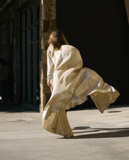 Adult female in flowing gown is walking toward a building with stone siding; sunlight and shadow accentuate textures and movement.