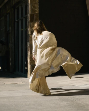 Adult female in flowing gown is walking toward a building with stone siding; sunlight and shadow accentuate textures and movement.