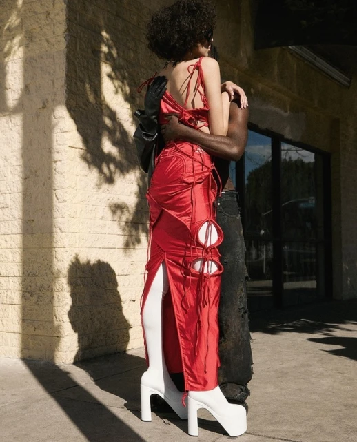 Woman in a red dress and platform boots embraced by a man outdoors in sunlight, creating shadows on the building's wall.