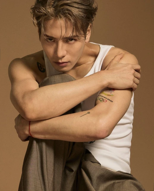 Portrait of a muscular adult male with dark hair, wearing a white tank top and grey pants, posing against a tan background.