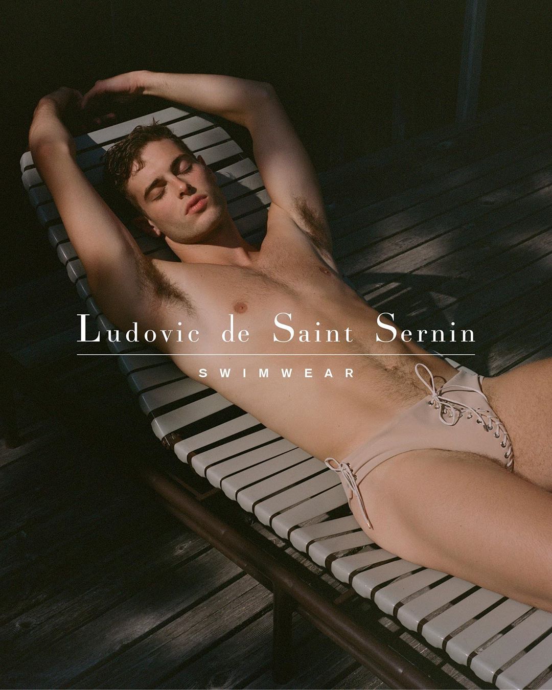 A light-skinned man poses on a lounger in neutral-toned swimwear, bathed in natural light. Features brand signage.