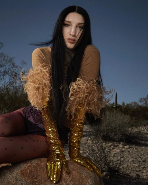 Portrait of an adult woman with long black hair wearing a tan turtleneck sweater, gold sequin gloves and patterned tights posing outdoors.