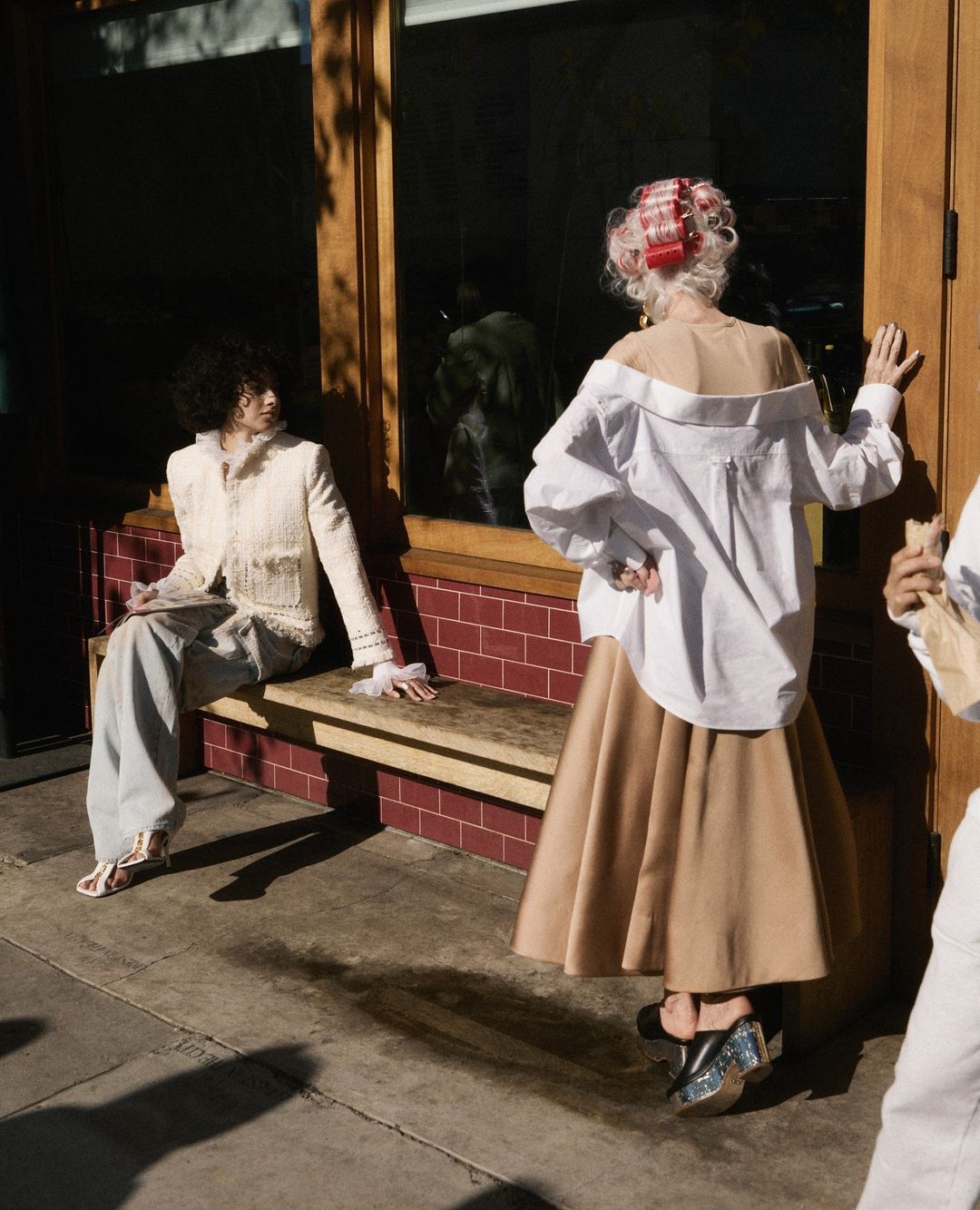 Two women in fashionable clothing pose in front of a brick building with glass windows on a sunny day, showcasing stylish outfits.