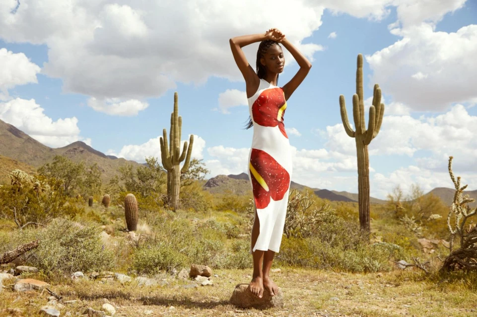 A young Black woman poses in a white floral-print dress in a sunny desert landscape with cacti, mountains, and cloudy skies, fashion shoot.