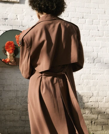Back view of a person with short curly hair wearing a brown trench coat standing before a white brick wall and circular mirror reflecting red flowers