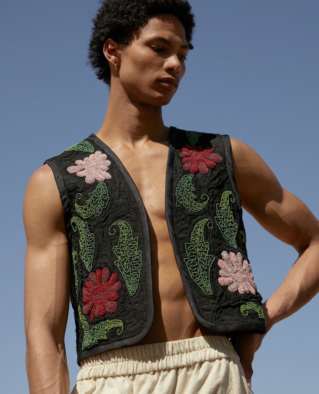 Man in embroidered vest poses in front of cloudless blue sky. He wears a sleeveless shirt and light-colored pants with hands on hips.