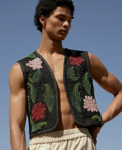 Man in embroidered vest poses in front of cloudless blue sky. He wears a sleeveless shirt and light-colored pants with hands on hips.