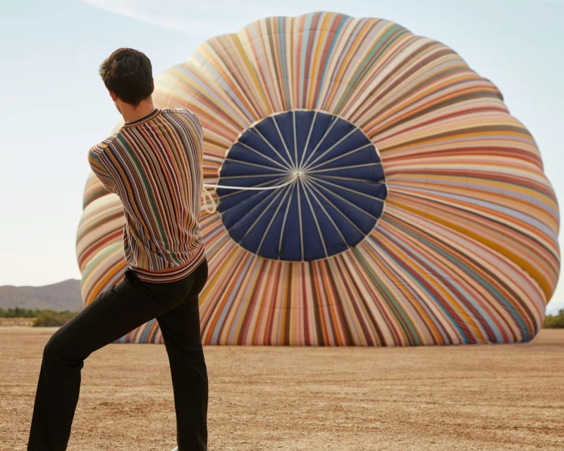 Adult male stands in a field with a deflated striped hot air balloon on the ground against a backdrop of hills and blue sky.