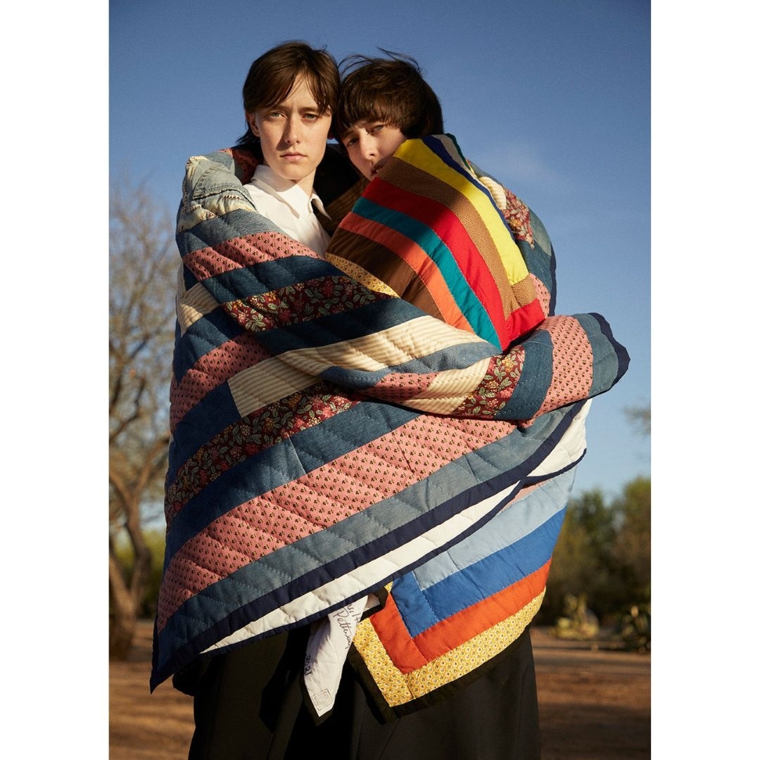 Two young adult women wrapped in a colourful patchwork quilt, standing together outdoors under a bright blue sky with desert vegetation.