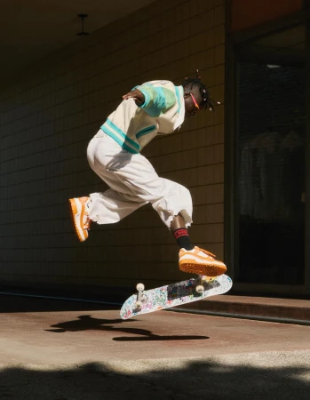 Athletic man mid-air performing a skateboarding trick in tan sneakers and white streetwear in an urban setting with shadows.