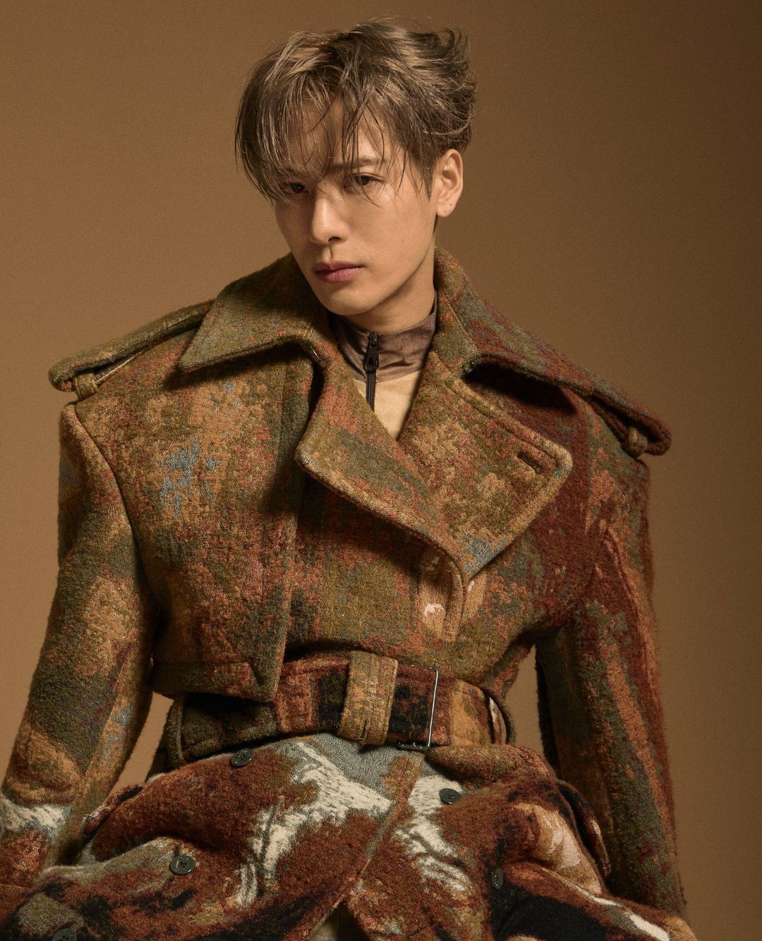 Adult male with light brown hair wearing a patterned brown jacket poses in a studio setting with neutral tan background. Fashion editorial.