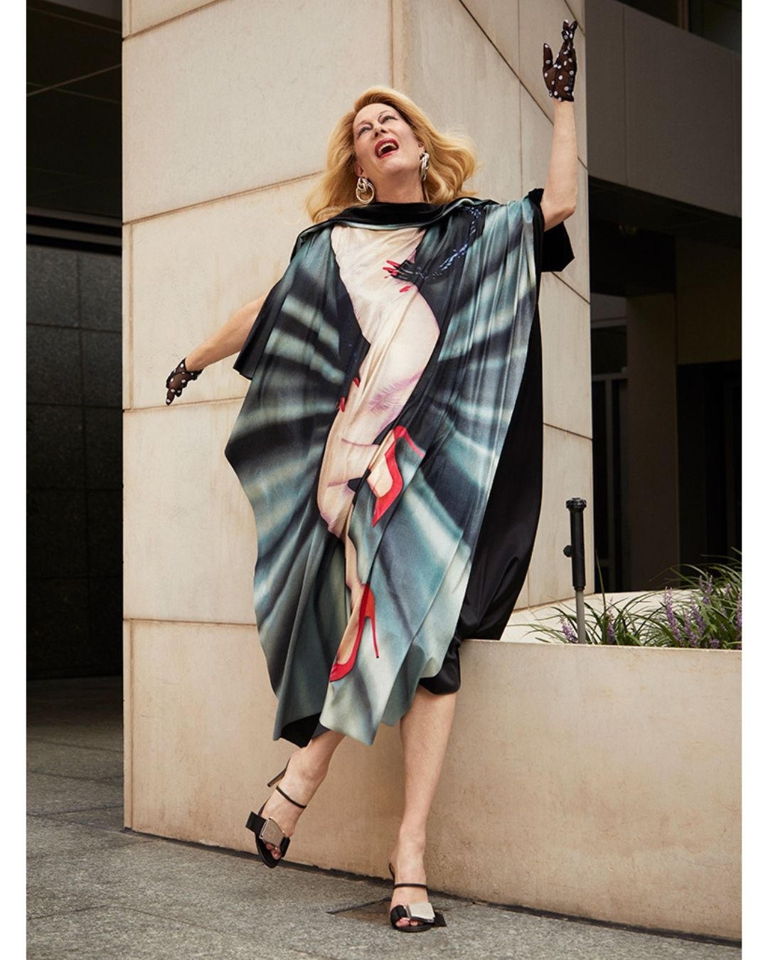Elegant adult woman in patterned kaftan poses on city street with raised arms, showing off black heeled sandals and spotted gloves.
