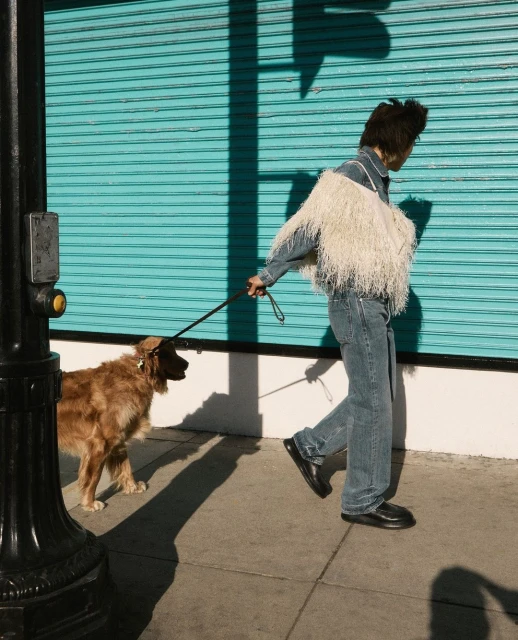 A person in a denim outfit walks a golden retriever dog on a leash on a sidewalk in front of a teal-colored building.