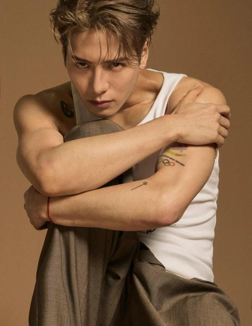 Portrait of a young adult male with light brown hair in a white tank top and grey pants sitting against a neutral tan backdrop.