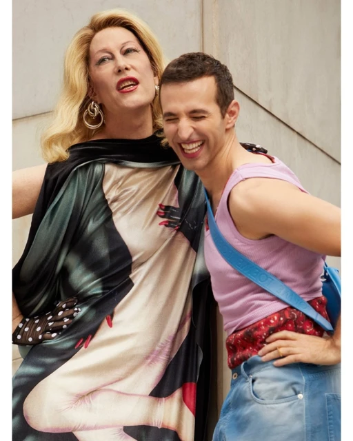 Two smiling adults pose indoors: a blonde person in a dress with a body print and a brunette person in a pink top and blue pants.