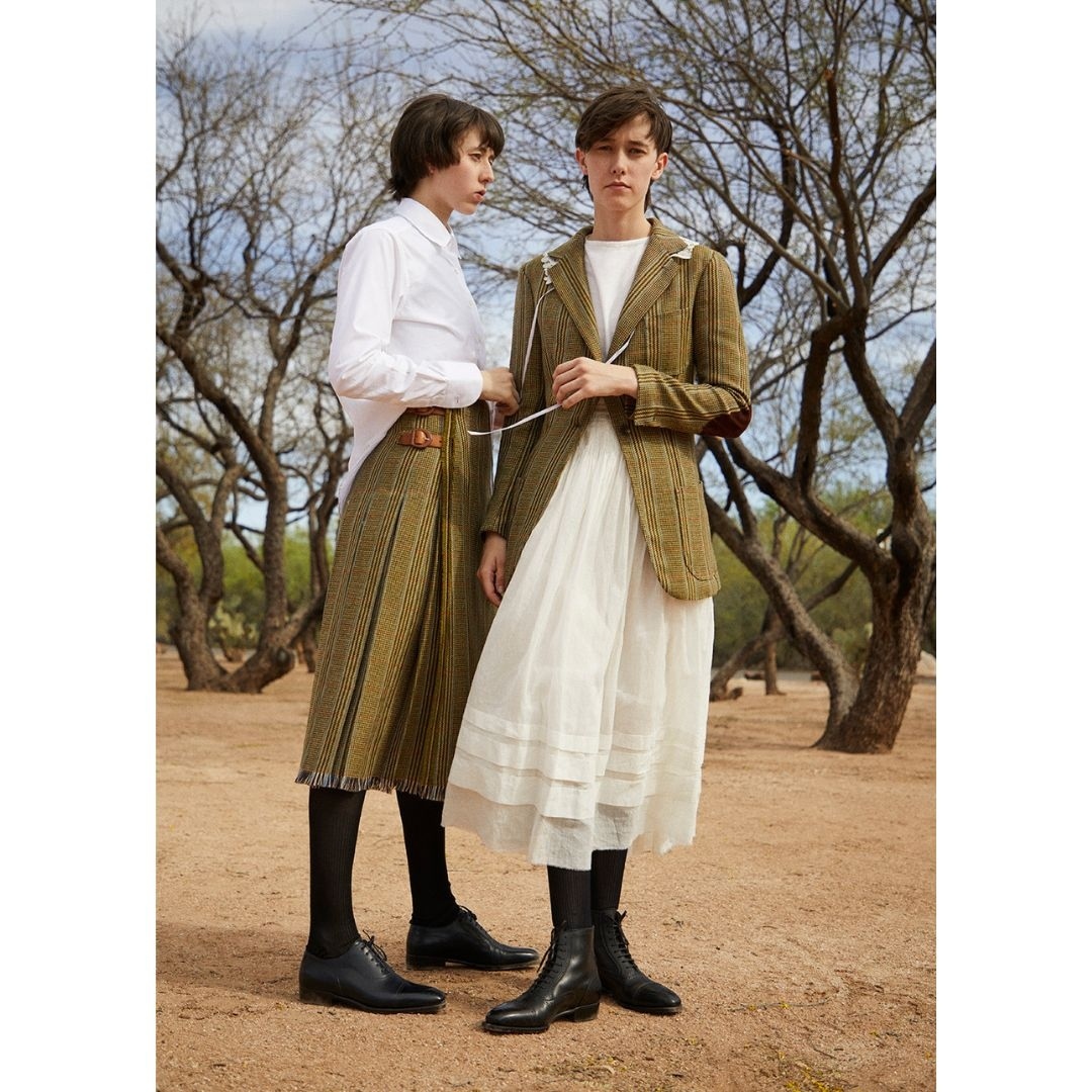 Two women in vintage-inspired fashion stand in a dry, sparse landscape featuring bare trees, both wearing skirts and blouses. Fashion editorial.