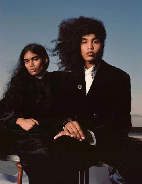 Two models pose in velvet jackets in front of a blue-grey sky. Dark hair, neutral makeup. A fashion shot in natural outdoor light.