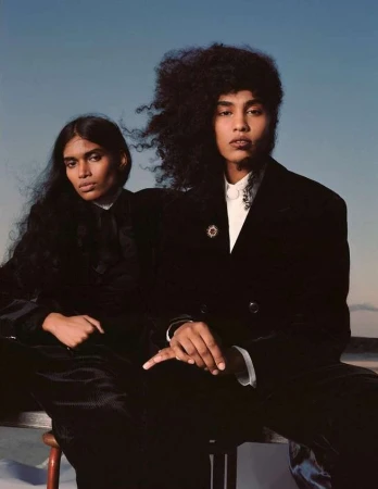 Two models pose in velvet jackets in front of a blue-grey sky. Dark hair, neutral makeup. A fashion shot in natural outdoor light.