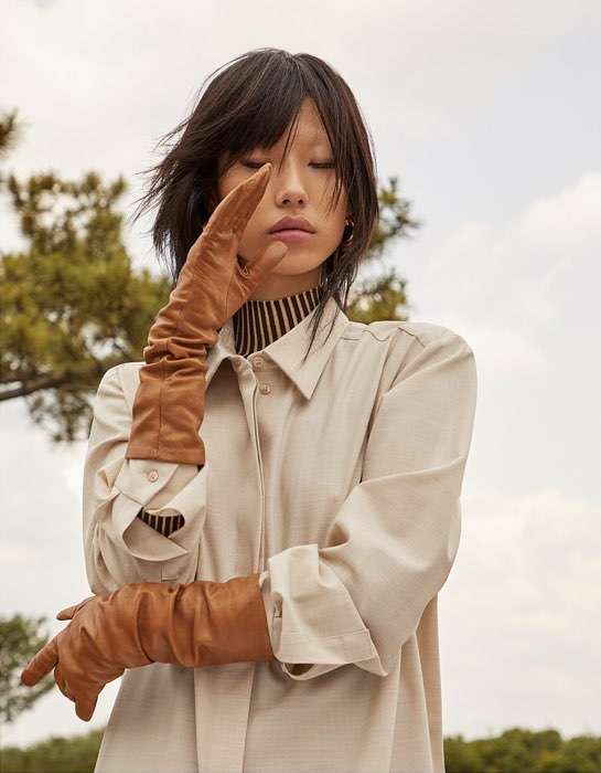 Fashion shot of a woman wearing long brown leather gloves and a light-colored shirt, posing outdoors with trees in the background.