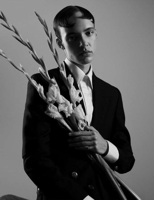 Monochromatic portrait of a person holding gladiolus flowers, wearing a suit and collared shirt against a neutral grey background.