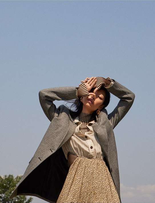 A stylish adult female model poses outdoors wearing a coat, patterned dress, and striped shirt against a clear sky backdrop in natural light.