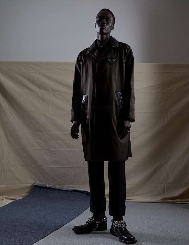 Full-length fashion portrait of an adult male wearing a dark overcoat and trousers with chain embellishments on his shoes, against a neutral backdrop.