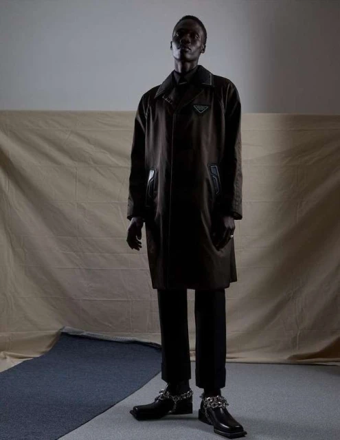 Full-length fashion portrait of an adult male wearing a dark overcoat and trousers with chain embellishments on his shoes, against a neutral backdrop.