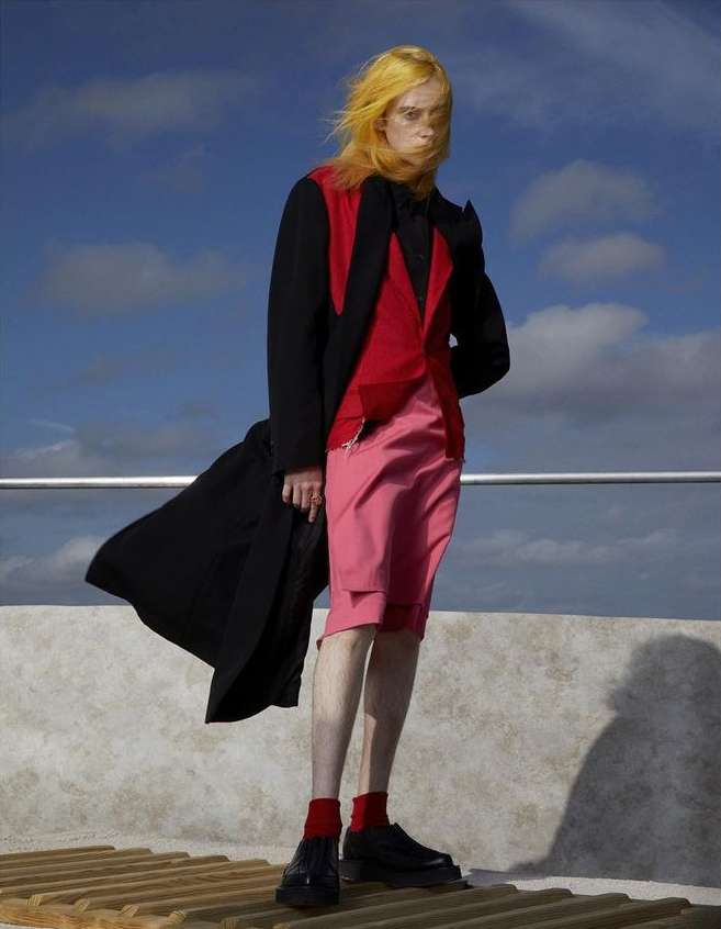 Fashionable figure posing outside on a rooftop, wearing a stylish red, black and pink outfit with matching yellow hair, and dark shoes.