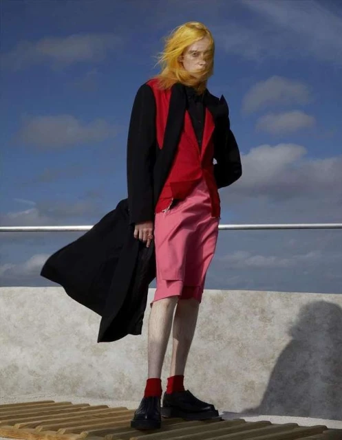 Fashionable figure posing outside on a rooftop, wearing a stylish red, black and pink outfit with matching yellow hair, and dark shoes.