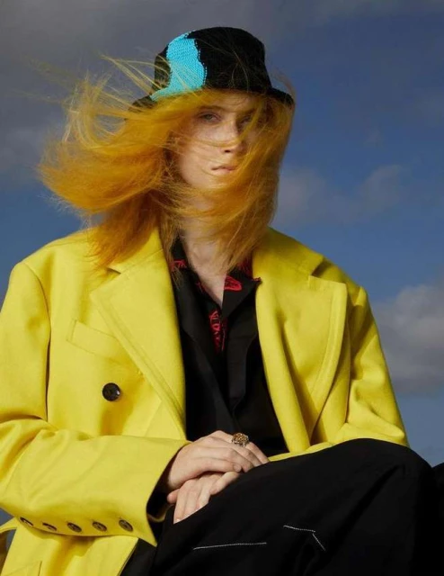 Portrait of a fair-skinned person with orange hair wearing a black and blue hat, yellow coat, and black shirt against a blue sky background.