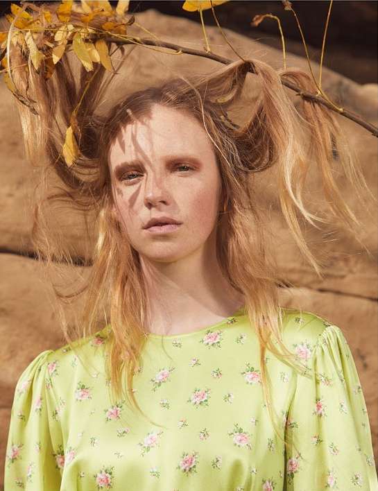 Portrait of a woman with blonde hair posing with leaves and branches; she wears a green floral print dress against a sandstone background.