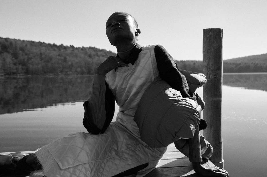 Monochrome portrait of a person relaxing on a wooden dock by a lake with forested hills in the distance on a sunny day.