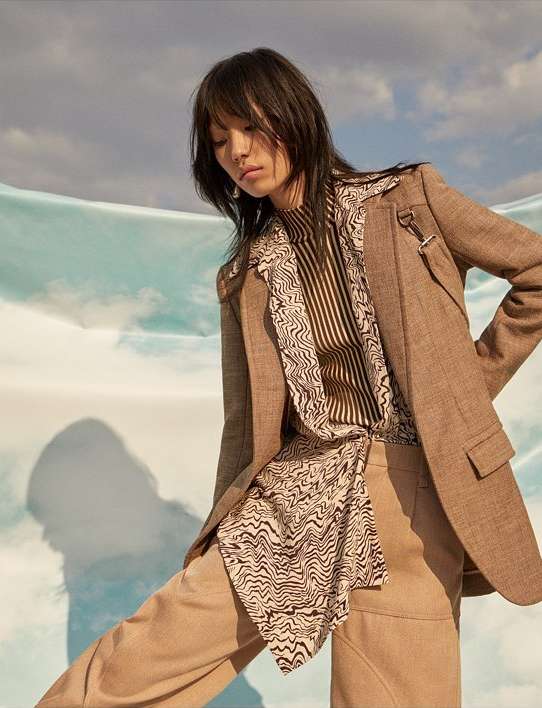 Brunette woman in a beige blazer and tan pants posing with a light blue backdrop. Fashionable, modern, and stylish.