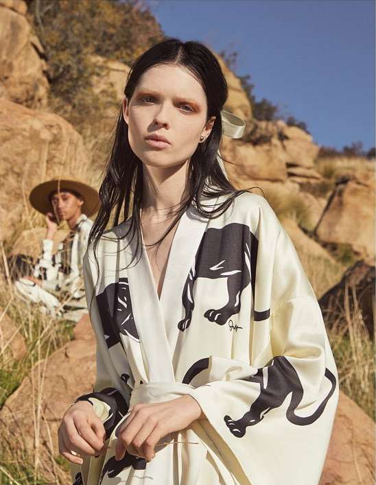 A woman wearing a black and white printed kimono-style robe poses outdoors near a seated person and rocky hills under a blue sky.