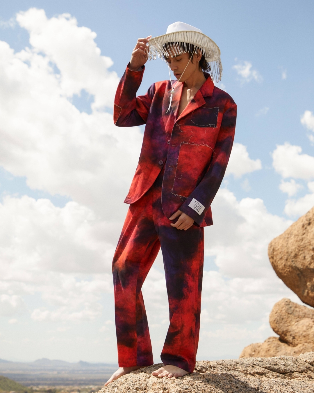 Adult male model in an artistic red and purple suit with white cowboy hat poses on a rock outcrop against a desert background and blue sky.