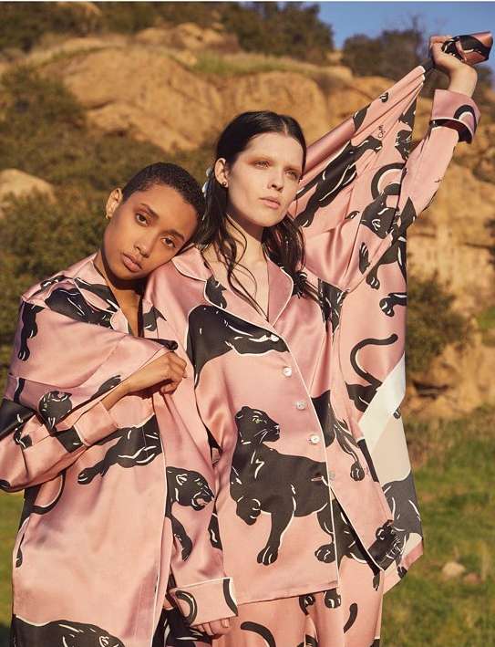 Two women model pink jaguar-print pajamas outdoors against a backdrop of hills and sky in a medium shot with natural light.
