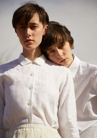 Two light-skinned adults in white collared shirts and neutral skirt. Close portrait with one resting head on the other's shoulder.