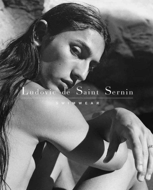Black and white close-up of a bare-chested adult with long hair, wearing swimwear, against a rocky background and swimwear logo.