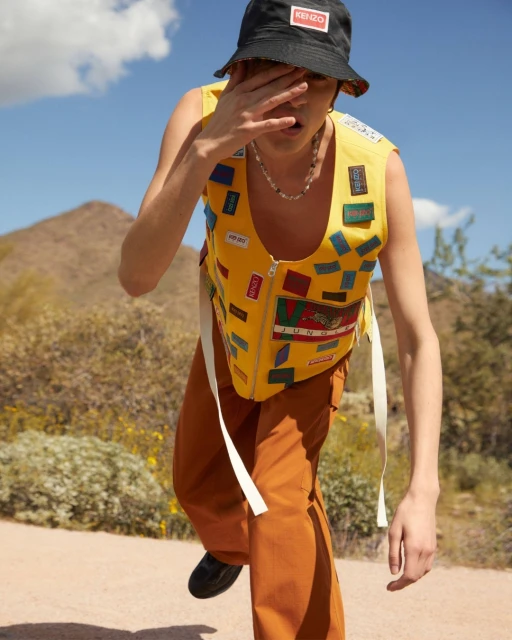 Male fashion model in yellow vest, rust pants, and black bucket hat in arid outdoor setting with mountains and blue sky.