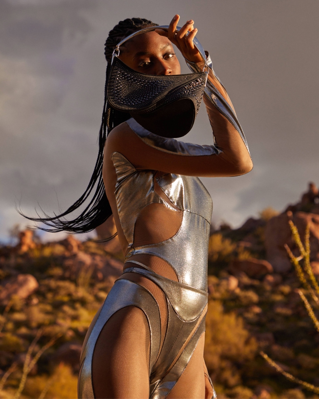 A young adult woman poses in a desert landscape, wearing a reflective silver bodysuit, and holding a sparkly black handbag over her face.
